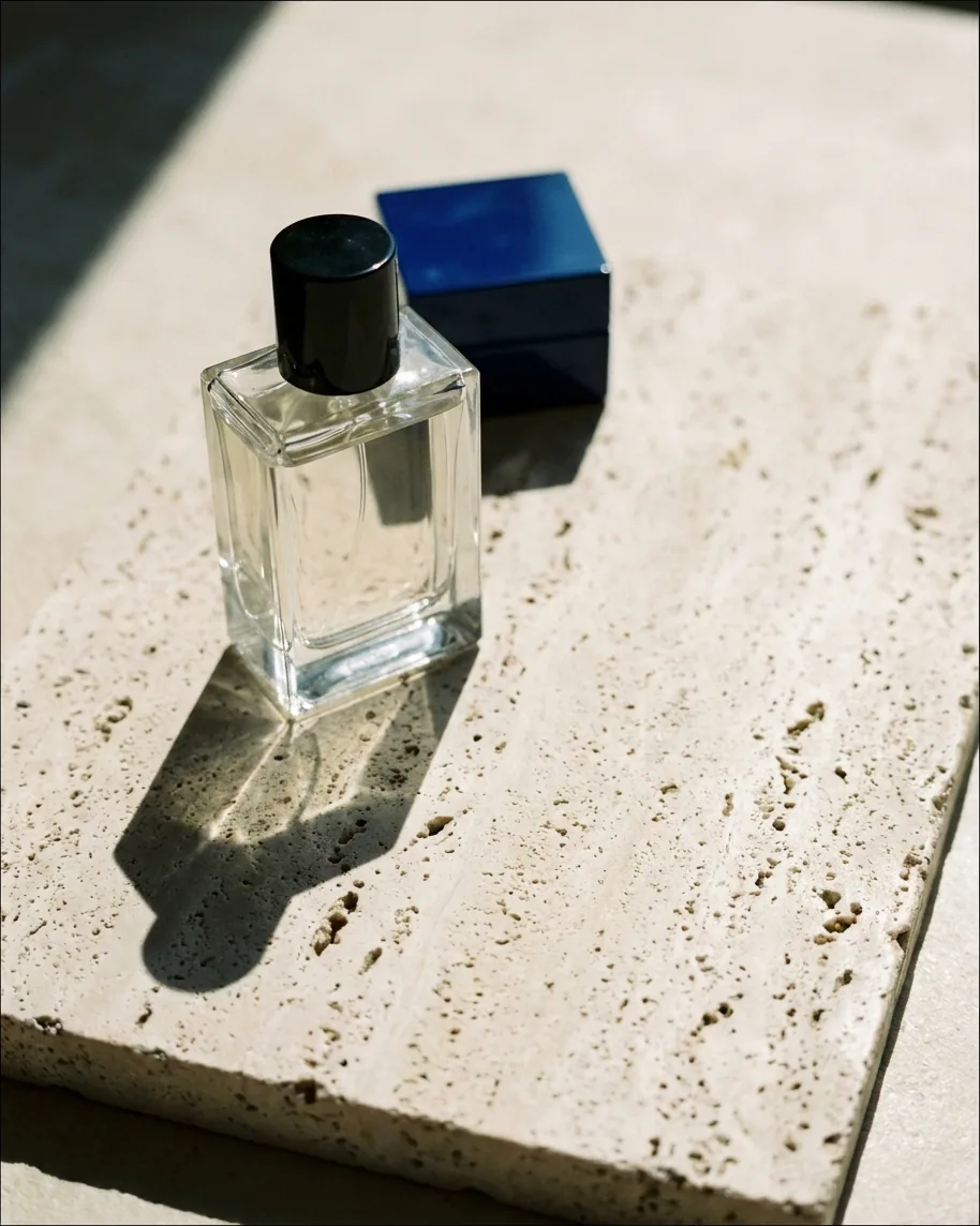 An unlabeled glass perfume bottle on raw travertine in hard noon light, a cobalt lacquer box behind it